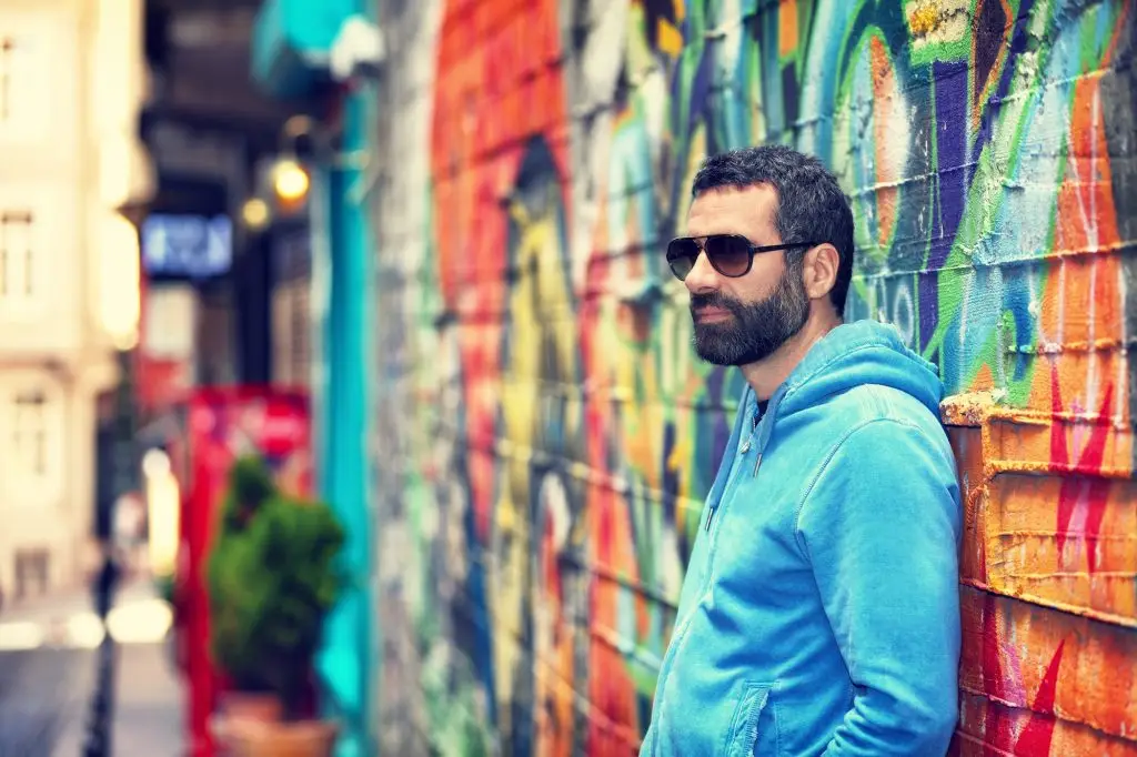 Bearded man in blue hoodie and sunglasses leaning against a colorful graffiti wall on a city street.