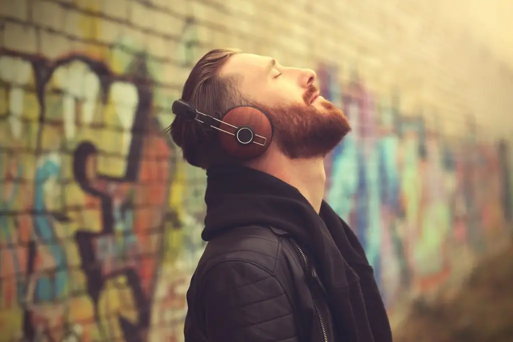 Bearded man with headphones, eyes closed, leaning against a colorful graffiti wall in a peaceful moment.