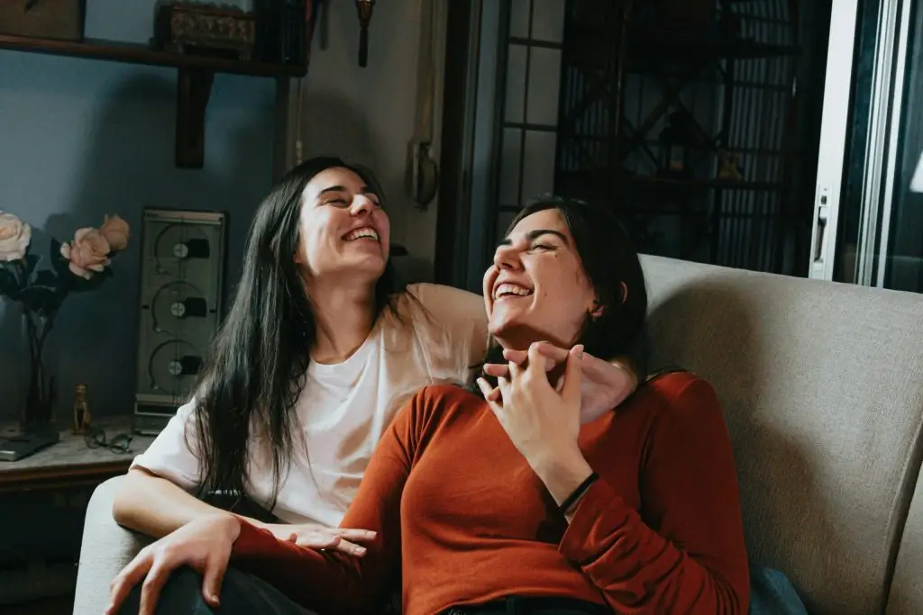 Two women laughing together on a couch, sharing a joyful, supportive moment.