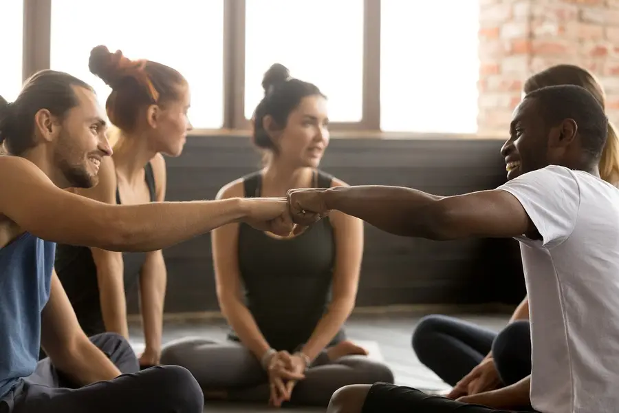 Two smiling men fist bump during a group therapy session as others sit nearby on the floor in a supportive circle.
