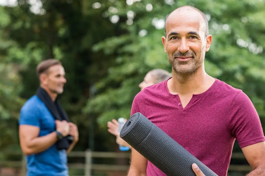 Smiling man holding a yoga mat outdoors with others nearby, ready for a holistic wellness session.