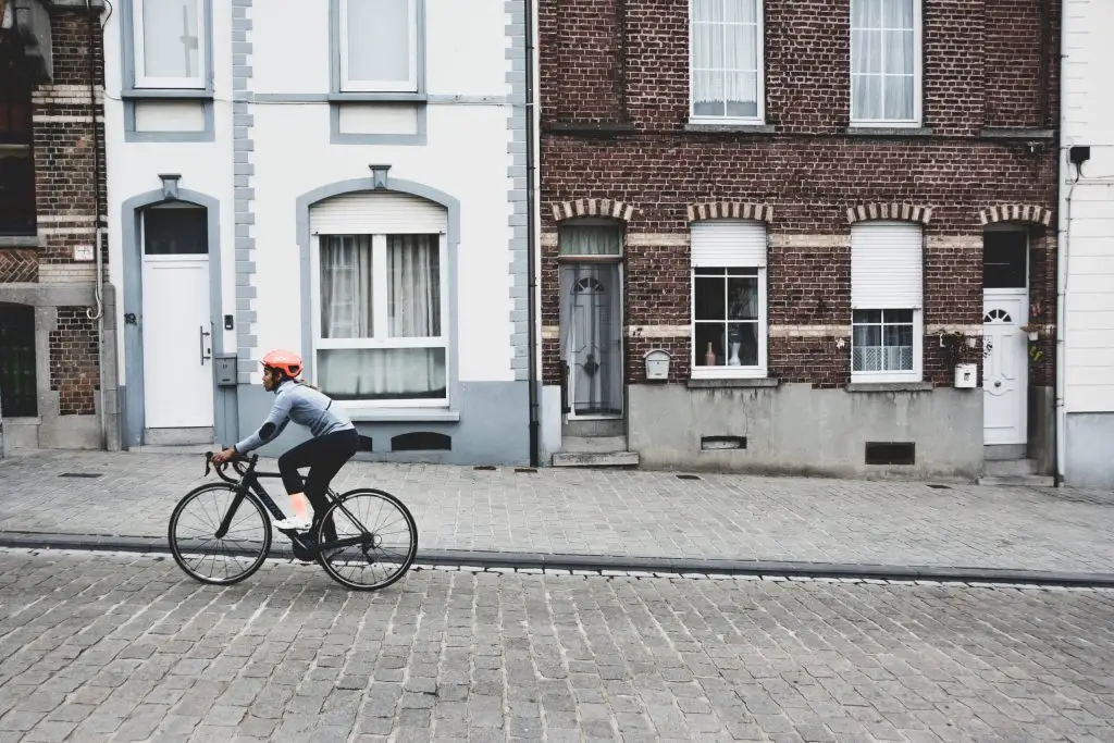 Cyclist in helmet rides a road bike along a cobblestone street past brick row houses.