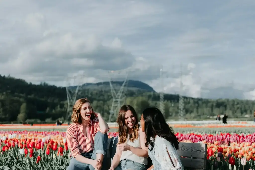 Three women laughing together while sitting on a bench in a colorful tulip field with mountains in the background.