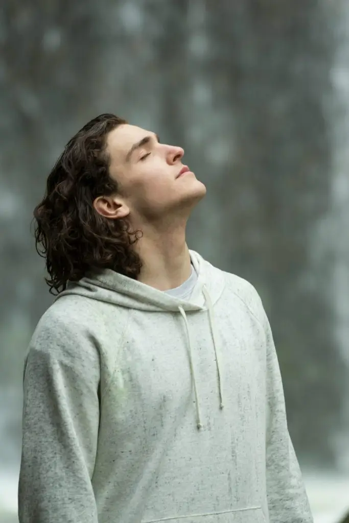 Young man in a gray hoodie with eyes closed and head tilted back, breathing calmly outdoors during recovery.