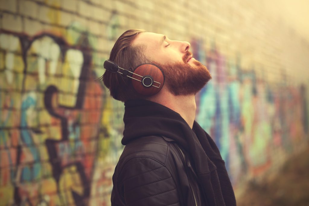 Bearded man with headphones and eyes closed leans against a colorful graffiti wall, finding peace in music.