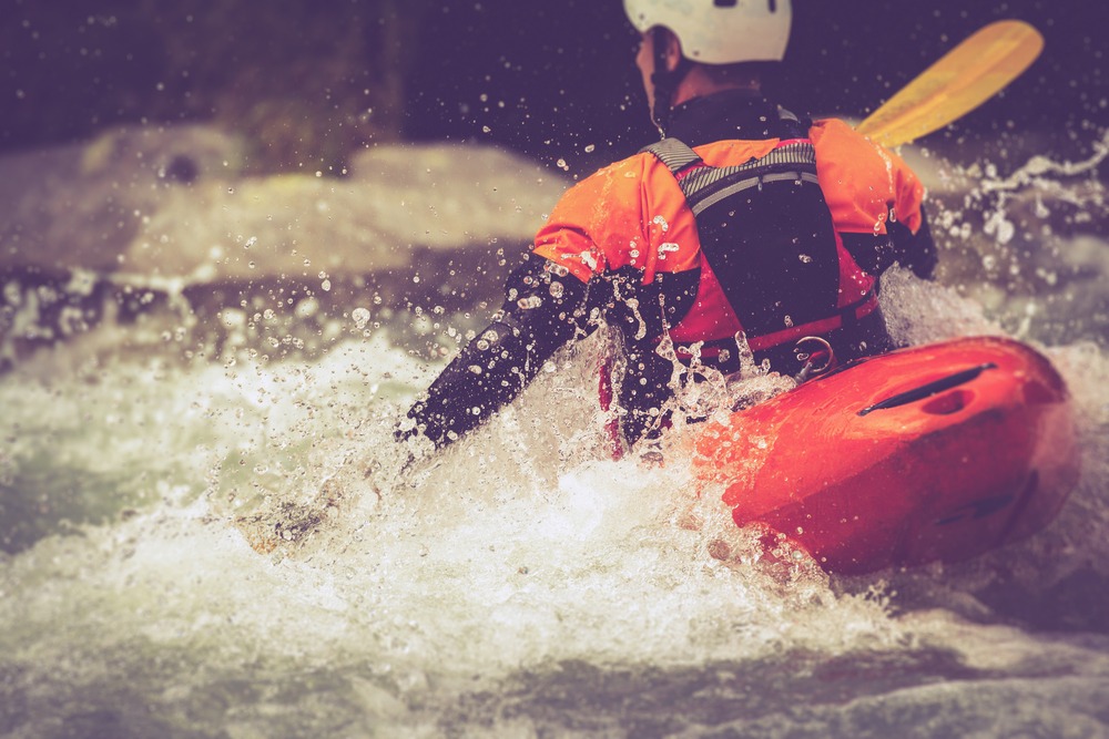 Kayaker in orange gear paddling through rushing whitewater rapids, water splashing all around.