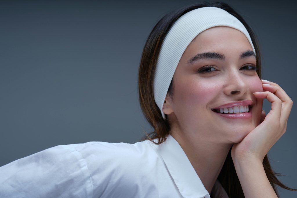 Smiling woman with white headband resting her chin on her hand against a gray background.