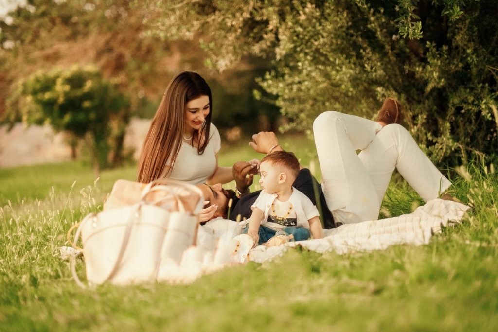 Family with young child enjoying a picnic together on a blanket in a sunny green park surrounded by trees.