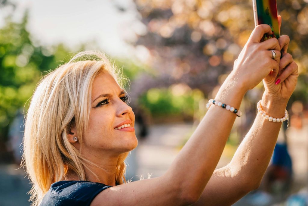 Smiling blonde woman holding up a phone outdoors in warm sunlight among blossoming trees.