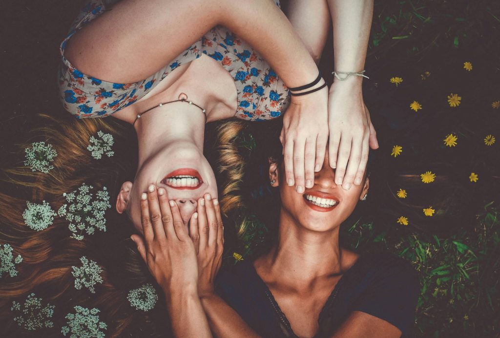 Two smiling young women lying head-to-head in grass and wildflowers, playfully covering each other's eyes.